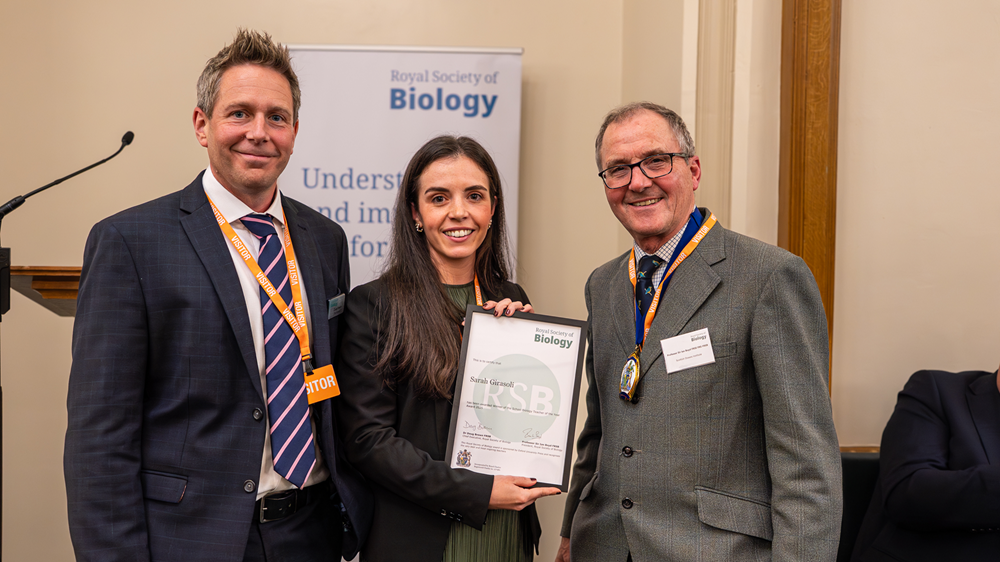RSB CEO Doug Brown, teacher Sarah Girasoli, and RSB President Ian Boyd standing side by side in front of RSB banner with Sarah holding certificate for School Biology Teacher of the Year 2025