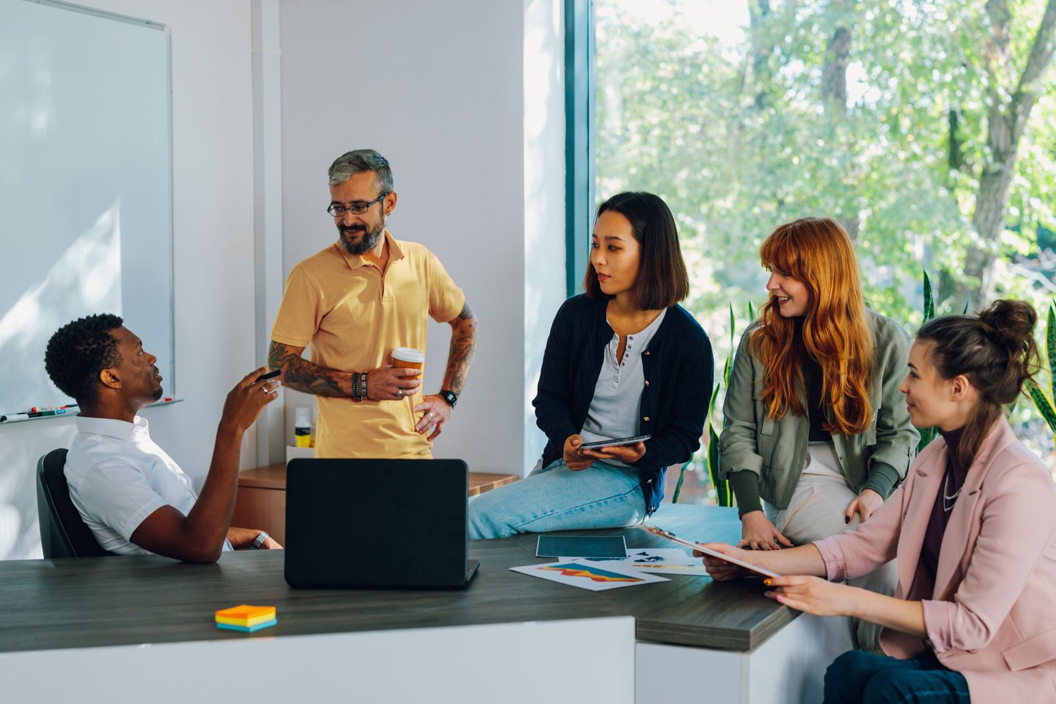 five people sitting and standing around a desk with a laptop on it